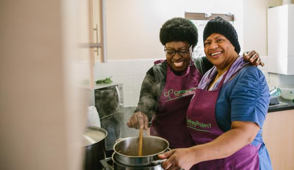 Two smiling women wearing purple Comfrey Project aprons are cooking together 