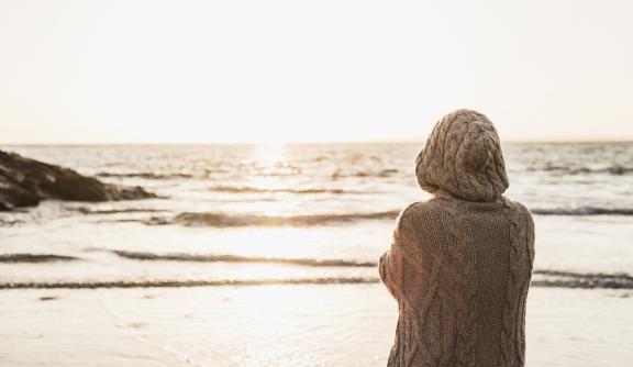 Woman with a hood and back to camera looks onto a beach