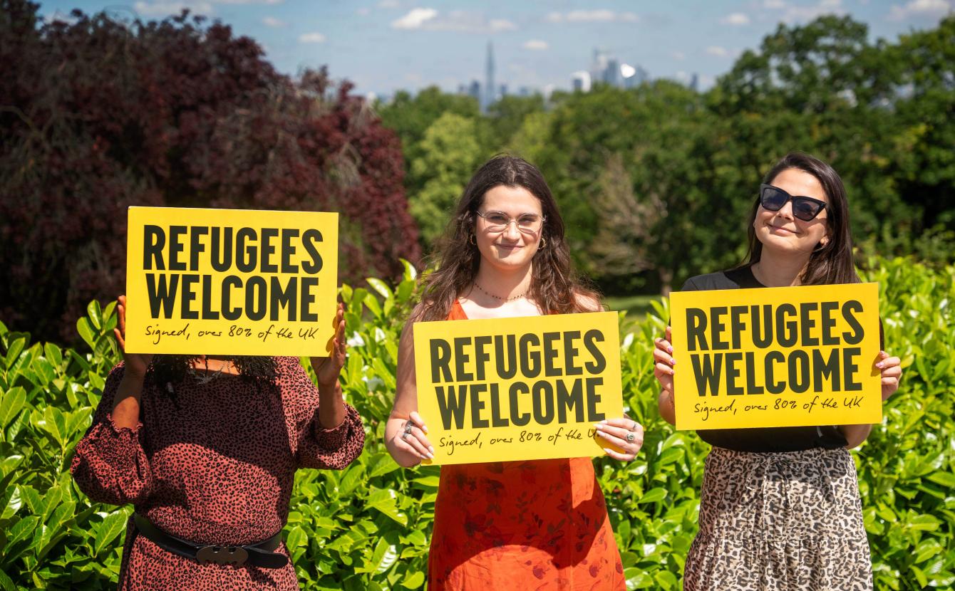 Three women holding a 'Refugees welcome' placard smiling at camera